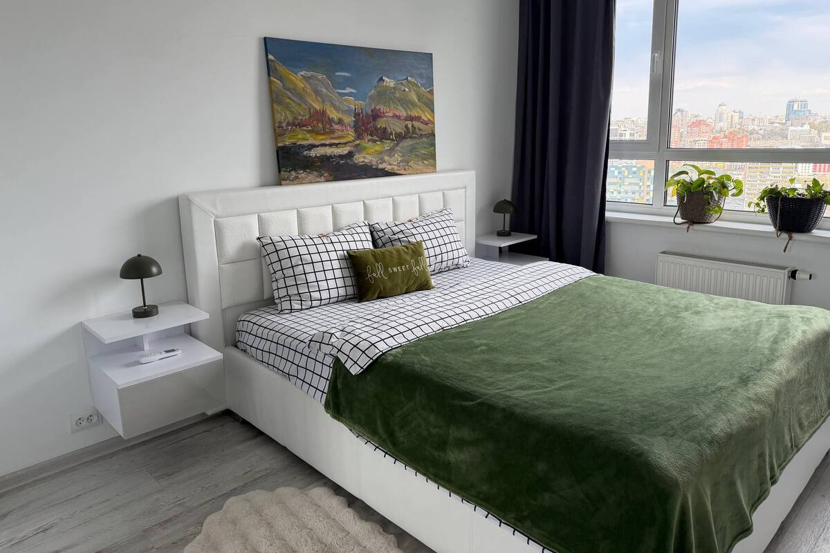 Un dormitorio moderno con vista a la ciudad, un cabecero blanco, una colcha verde y dos mesitas de noche con lámparas.