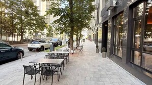 Un espace extérieur aménagé avec une terrasse dallée, des tables et des chaises, un trottoir, des voitures garées et un bâtiment doté de grandes fenêtres.
