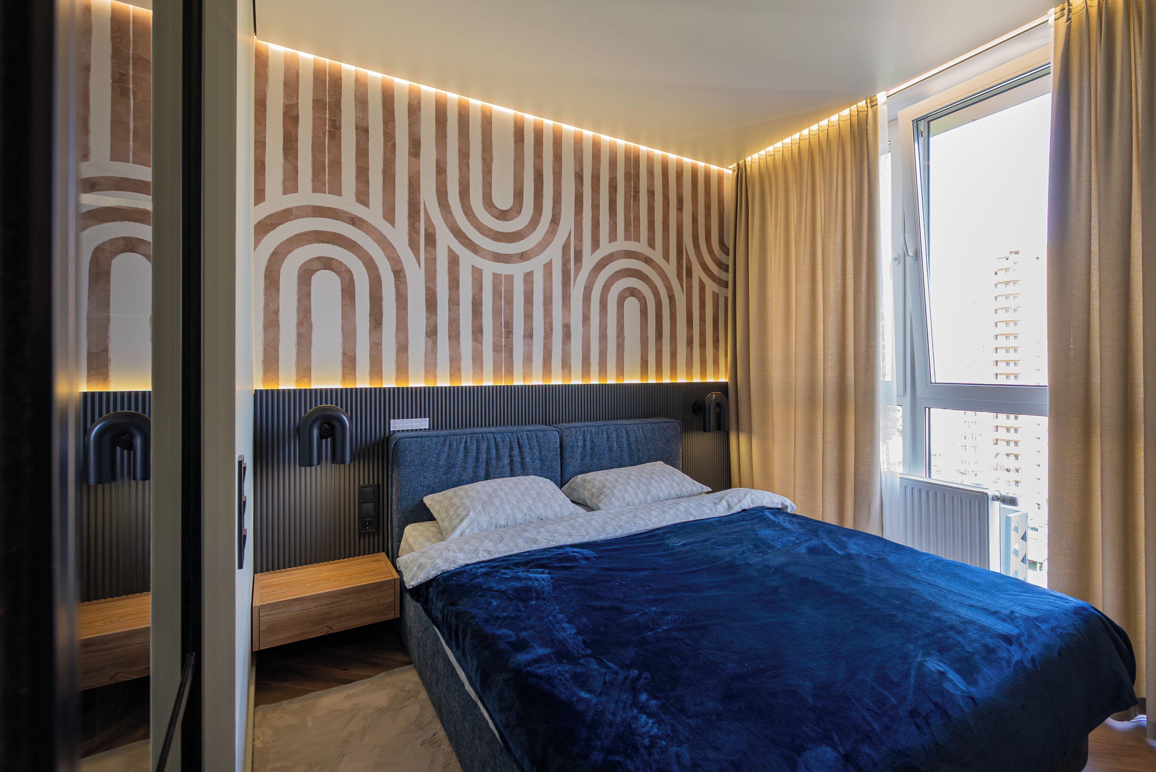 A modern bedroom with a large bed, a headboard with built-in lighting, a window with curtains, and a wall with a geometric pattern.