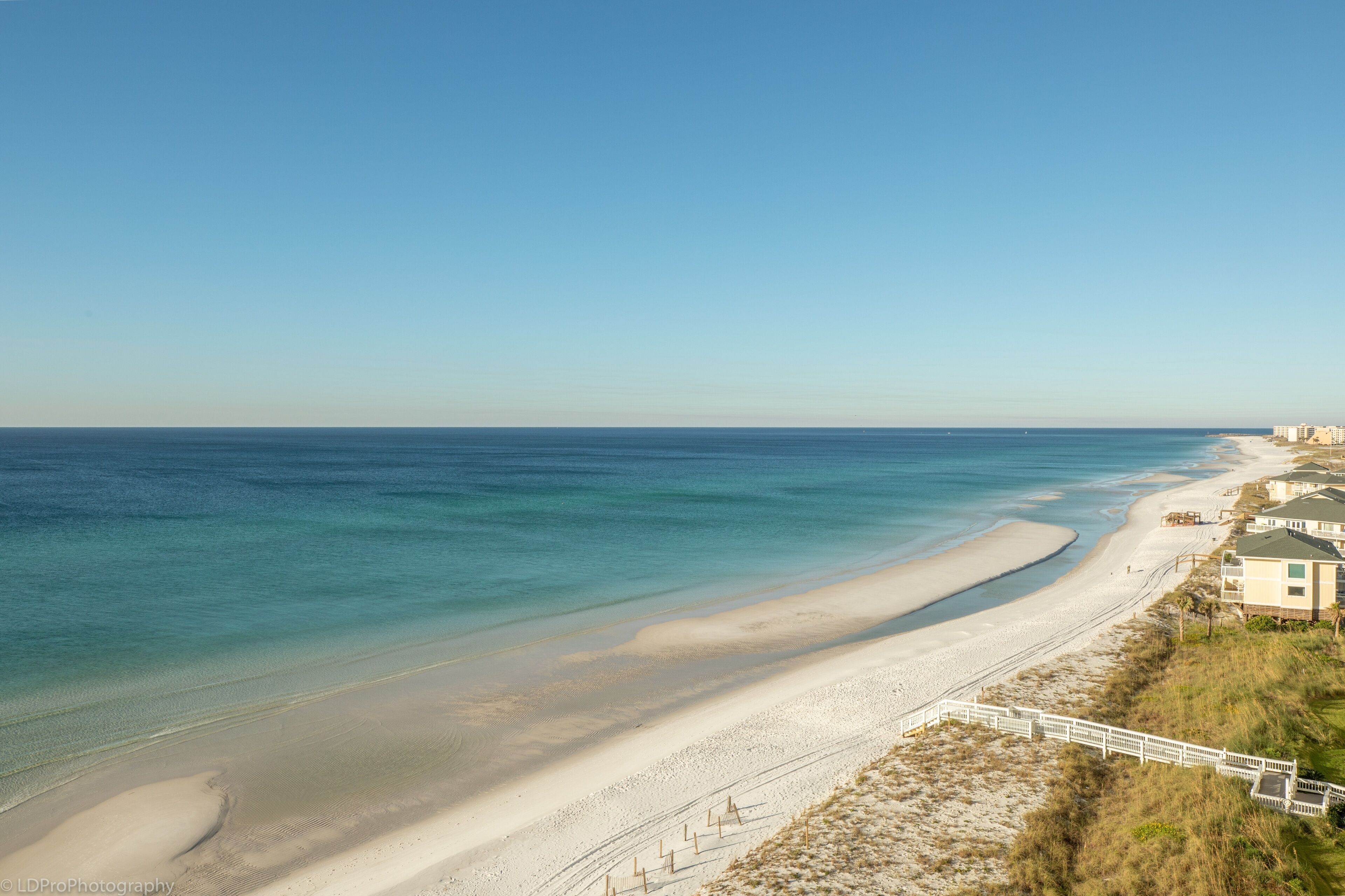 A coastal view with a sandy beach, a white fence, and a building with a flat roof.