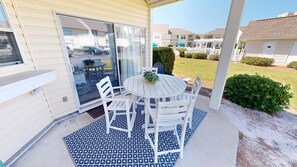 Un patio techado con una mesa redonda y sillas blancas, una puerta corrediza de vidrio y vistas a un barrio residencial.