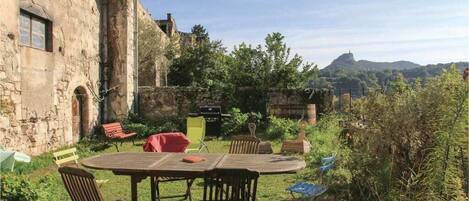 A rustic outdoor dining area with a round table and chairs, surrounded by greenery and a stone building.