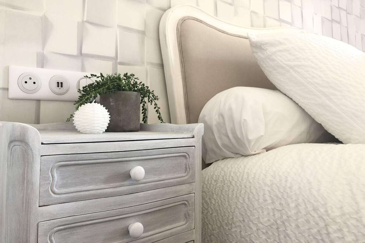 Uma cama bem arrumada com cobertura branca texturizada, uma mesa de cabeceira com uma planta em vaso e uma parede com padrão geométrico.