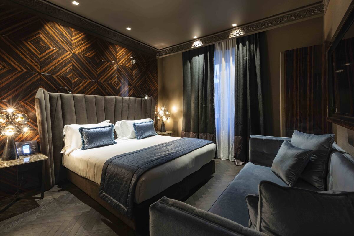 A modern hotel room with a large bed, a sofa, and a nightstand with a lamp.