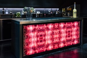 Zona bar con un pannello illuminato di rosso, bottiglie, bicchieri e un vaso con fiori.
