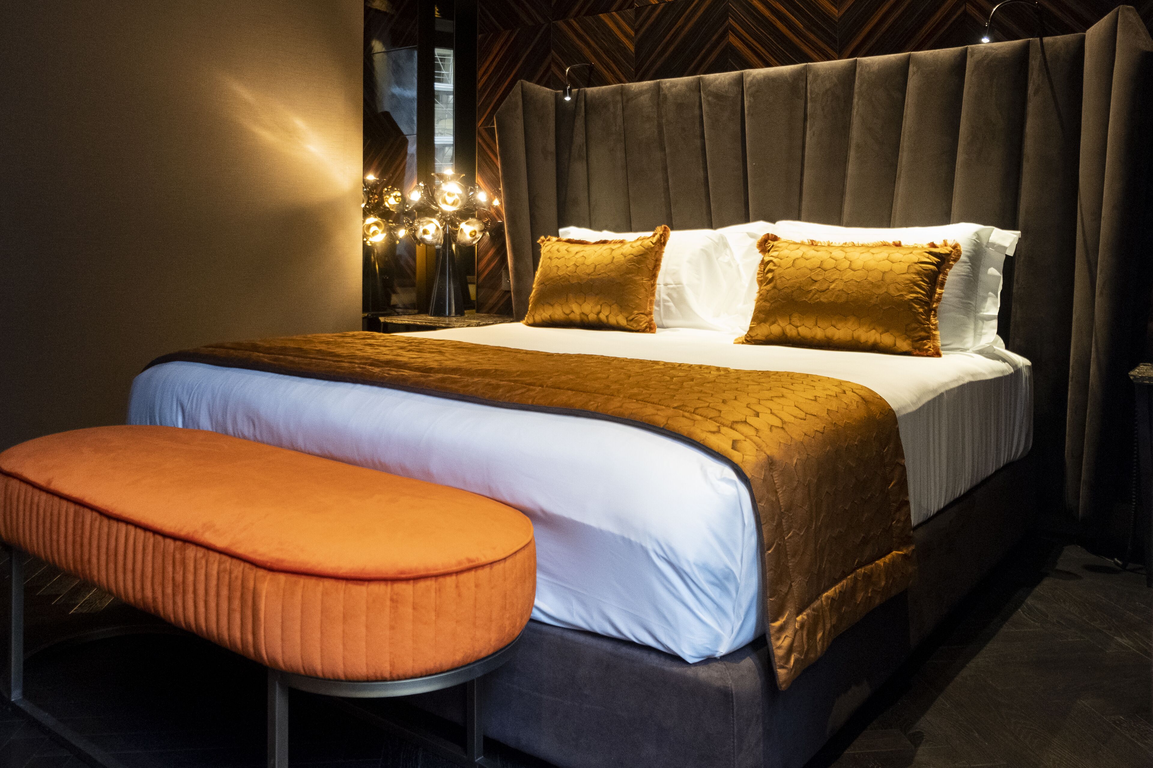 Una cama grande con cabecera capitonada, un banco al pie de la cama y una mesita de noche con una lámpara.