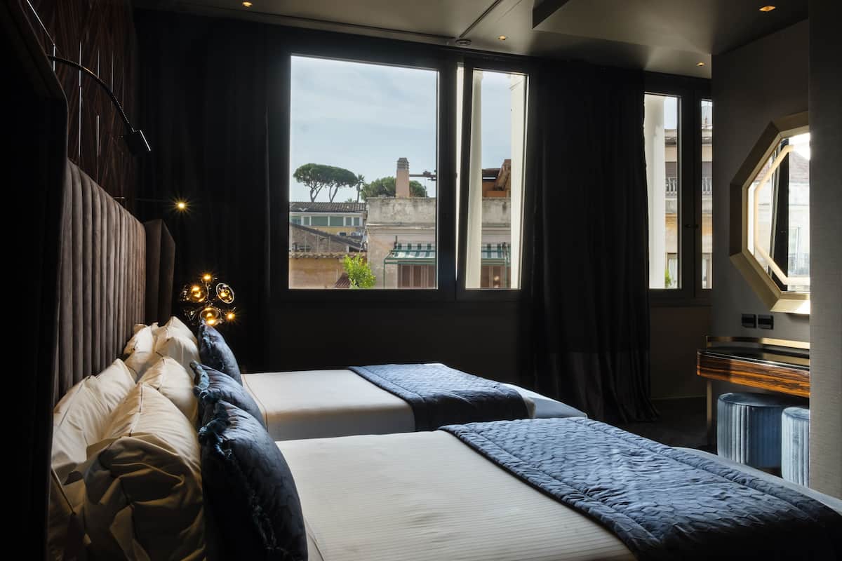 A hotel room with two beds, a large window with a view of buildings, a desk with a lamp, and a small window on the right.