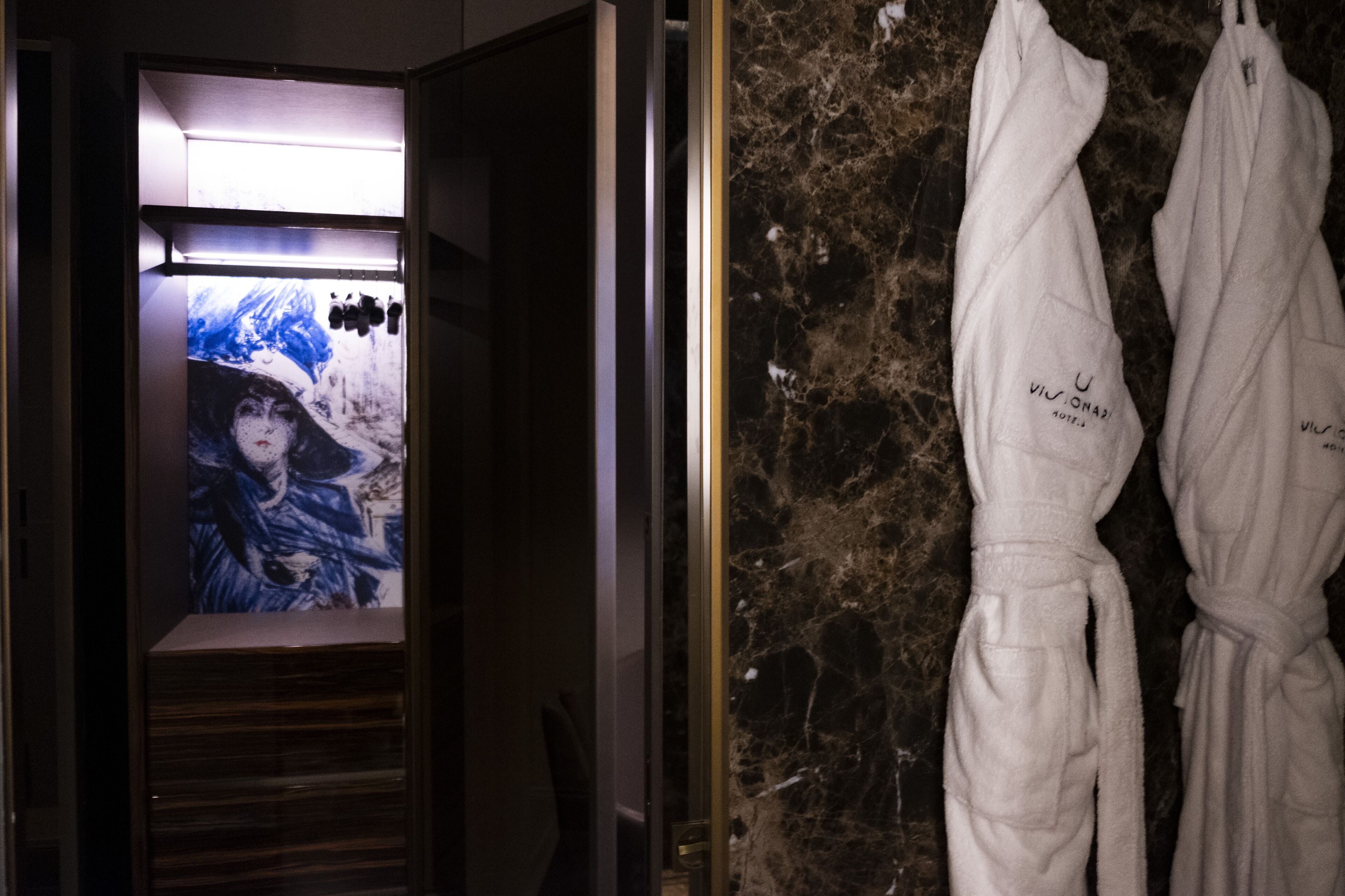 A bathroom with a marble wall, a shower area, and two white robes hanging on hooks.