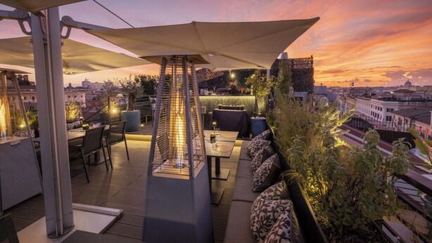 An outdoor terrace with tables and chairs, a large umbrella, and a city view at sunset. - U-Visionary Roma Hotel (Rome)
