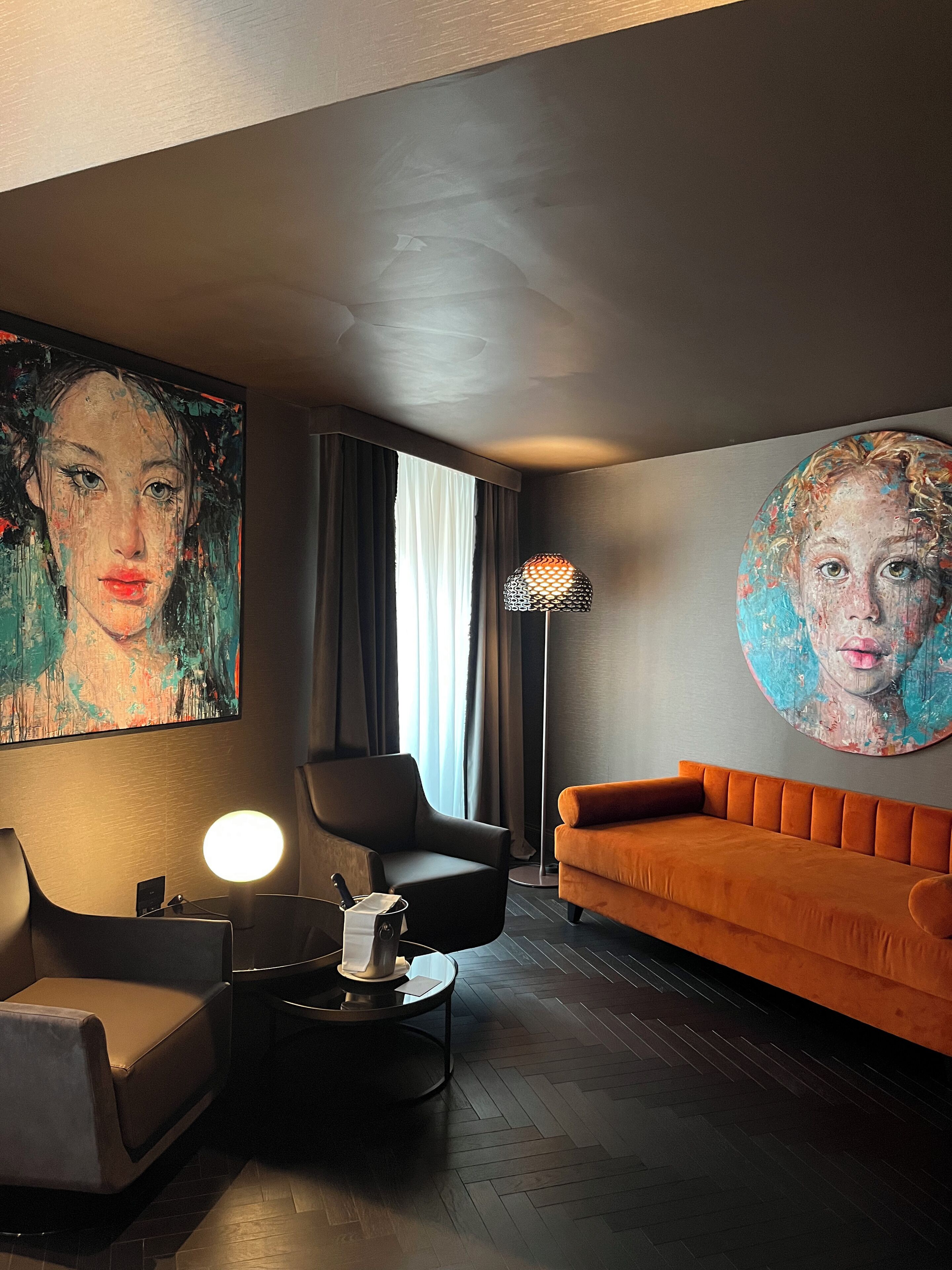 Ein modernes Wohnzimmer mit dunklem Holzboden, einem runden orangefarbenen Sofa, einem schwarzen Sessel, einem Couchtisch mit Lampe und zwei großen abstrakten Bildern an den Wänden.
