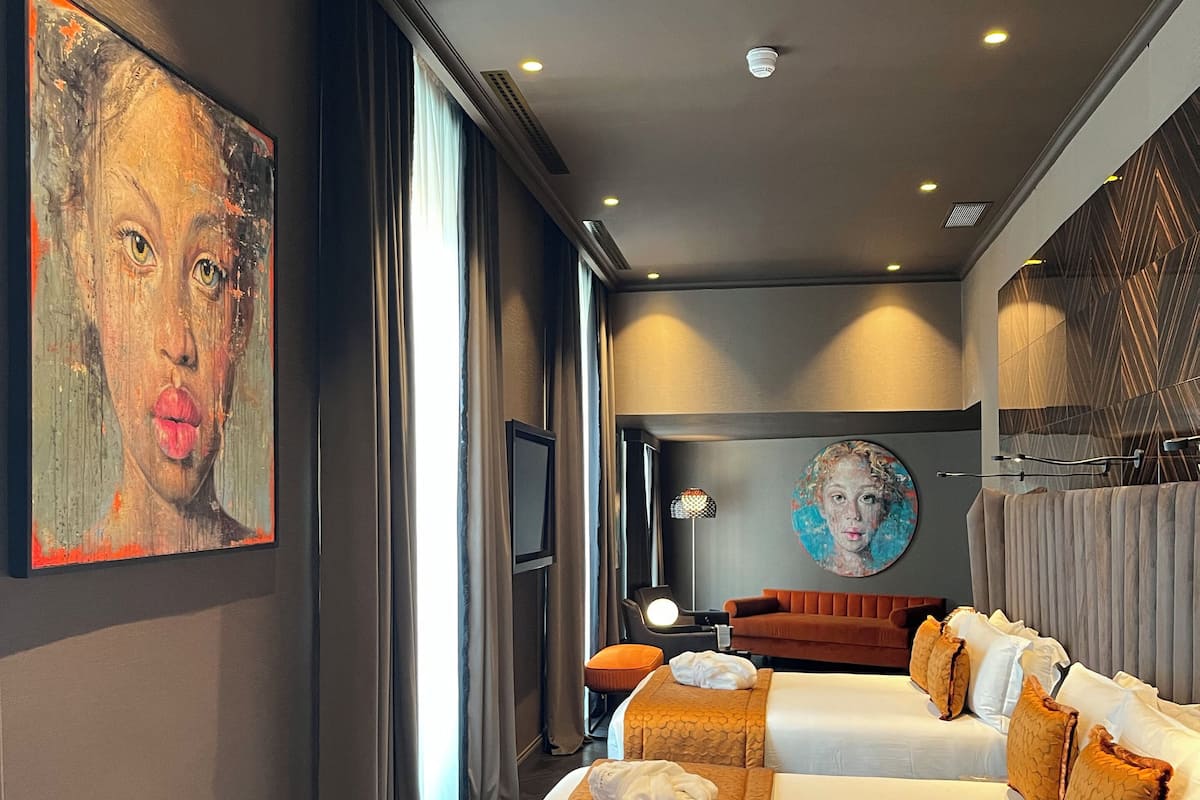 A hotel room with two beds, a large painting on the wall, a modern lamp, and a view of the city through the window.