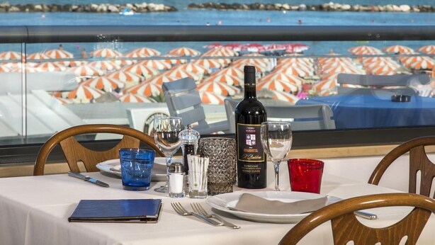 A table set with a bottle of wine, glasses, and a menu, overlooking a beach with umbrellas. - Hotel Igea Spiaggia (Bellaria-Igea Marina)