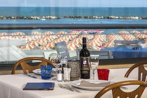 Una mesa con una botella de vino, copas y un menú, con vistas a una playa con sombrillas.