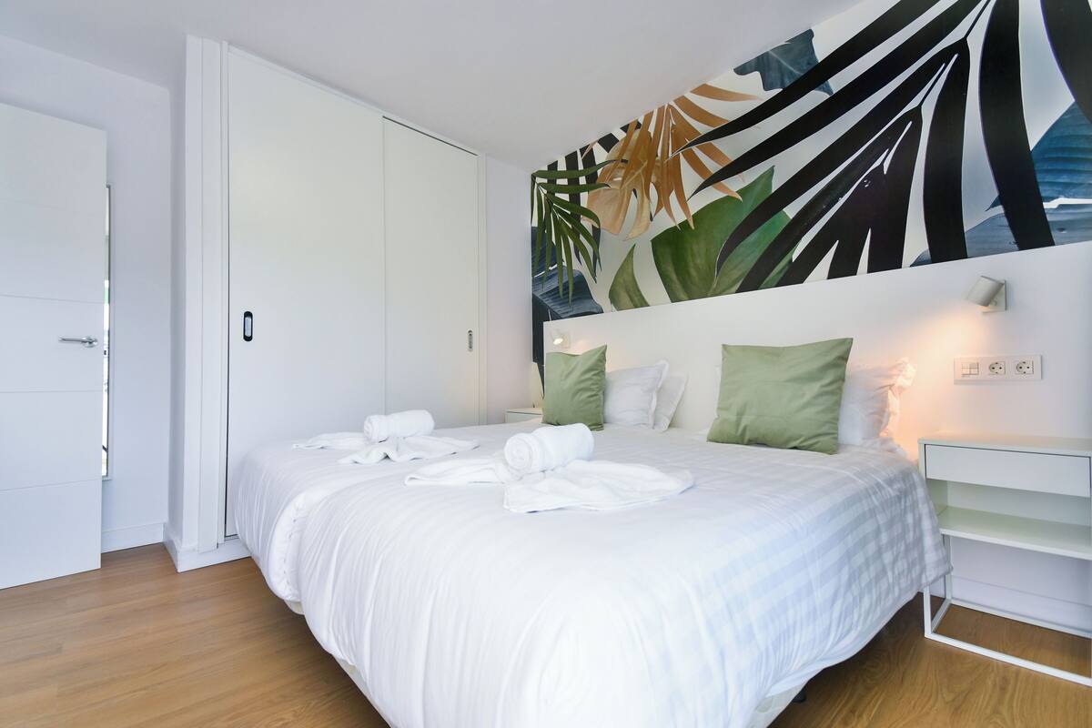 Un llit ben arrenjat amb roba blanca i tovalloles enrotllades, una paret amb mural de fulles tropicals i una taula de nit blanca amb una làmpada.