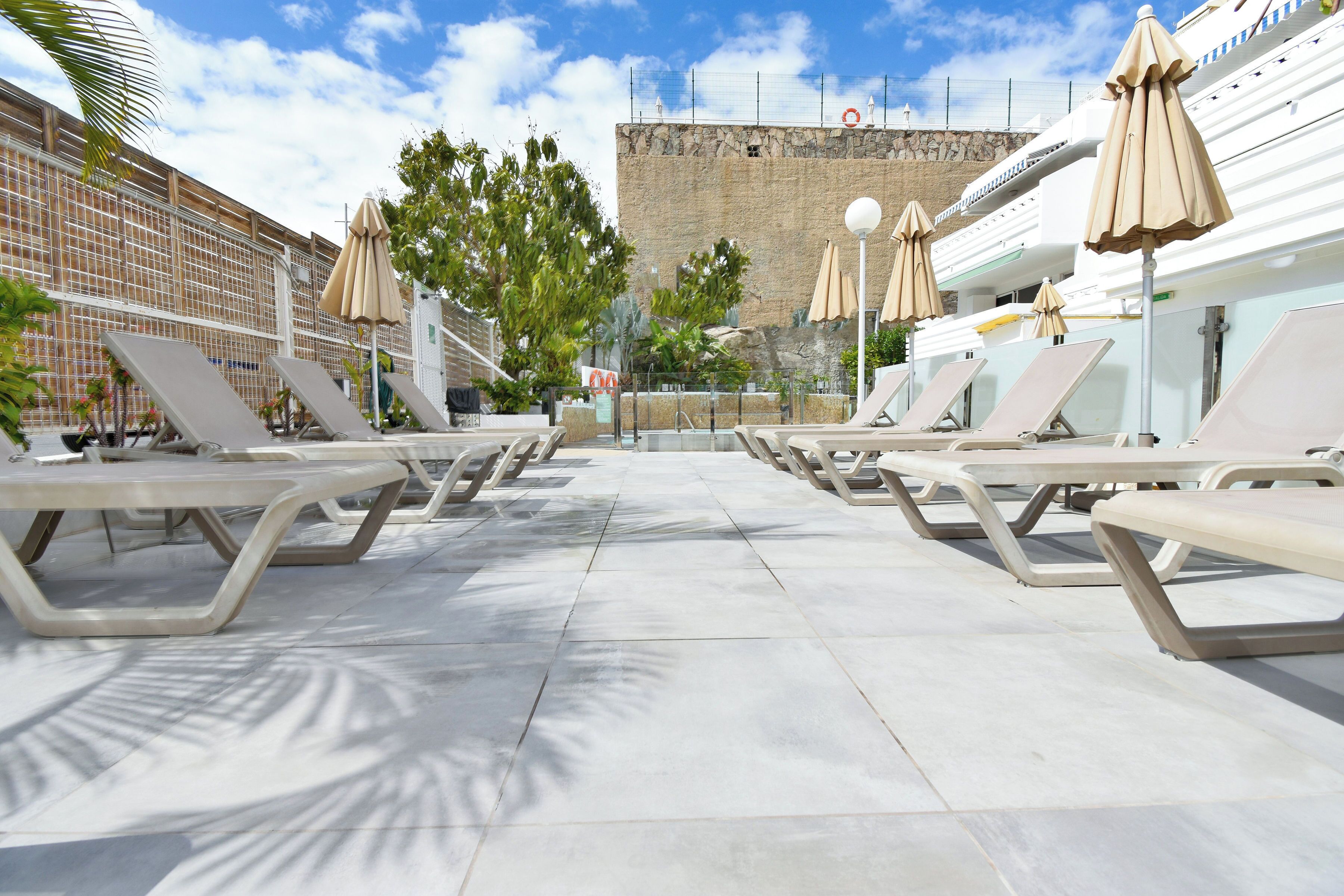 Zona de piscina con tumbonas y sombrillas, una pared de piedra al fondo y cielo despejado.
