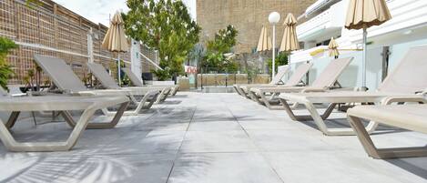 Zona de piscina con tumbonas y sombrillas, una pared de piedra al fondo y cielo despejado.