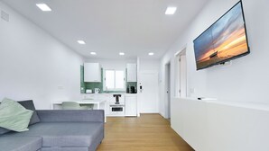 A modern living room with a grey sectional sofa, a flat-screen TV mounted on the wall, and a kitchen area in the background. - Palm Suites (Mogan)