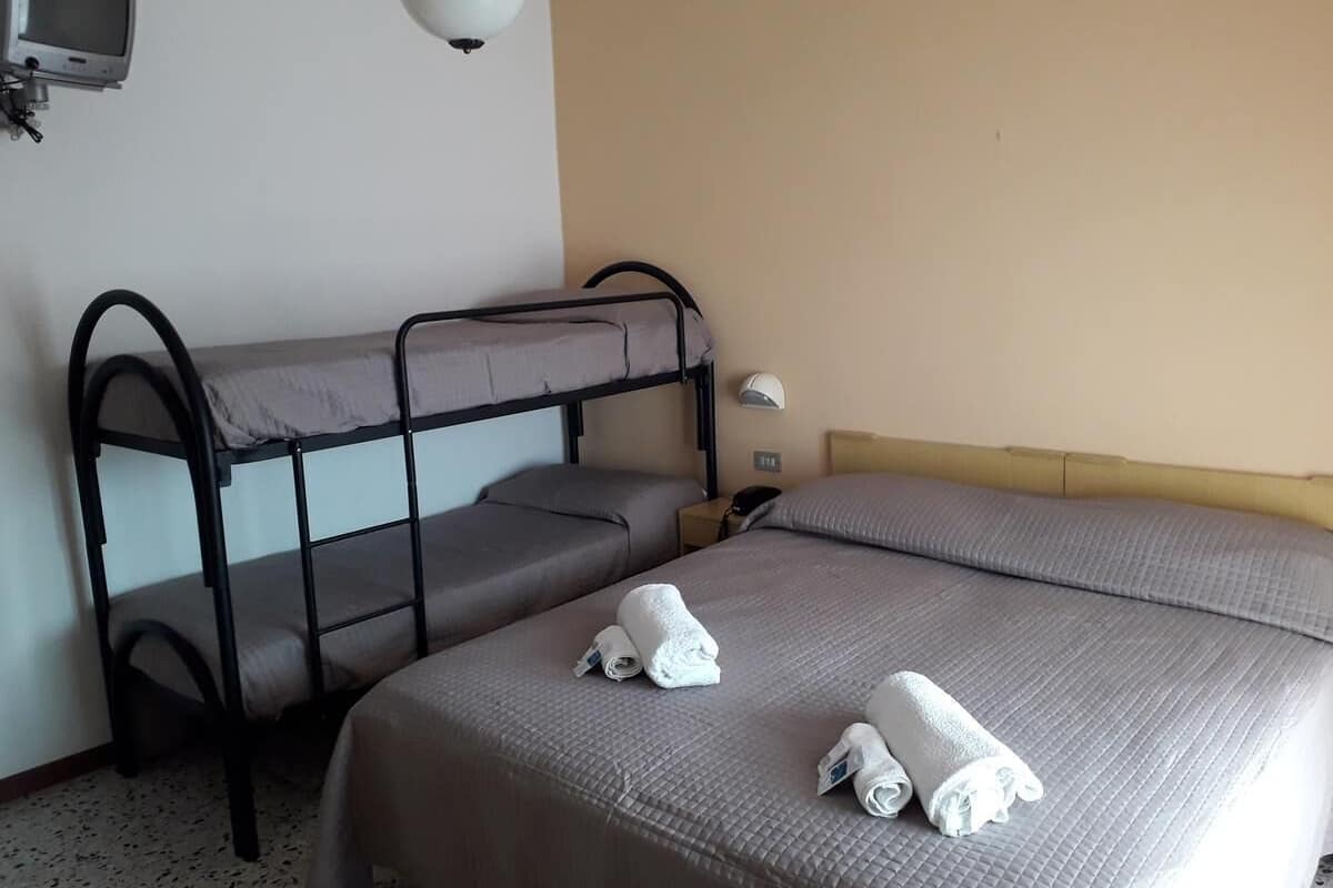 Een kamer met stapelbedden en een enkel bed, allebei met grijze beddengoed. Er staat een kleine tafel met een lamp en een telefoon.