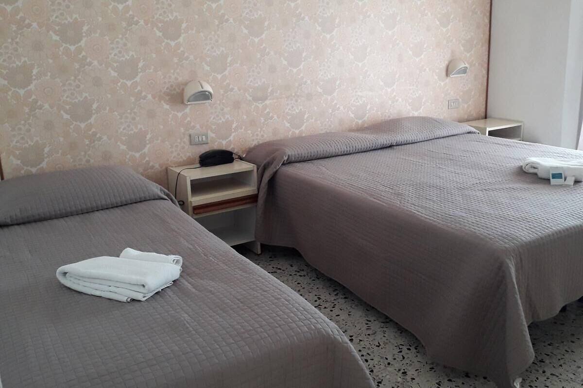 Een hotelkamer met twee bedden, elk met een grijze plaid en een witte handdoek erop. Daartussen staat een klein houten nachtkastje met een lamp.