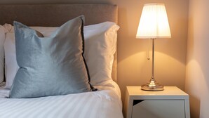A neatly made bed with white linens and two pillows, a bedside table with a lamp, and a small drawer. - Elliot Suite No3 - Donnini Apartments (Ayr)