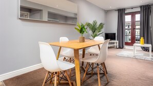 A modern living room with a wooden dining table, white chairs, a TV, and a large mirror. - Elliot Suite No3 - Donnini Apartments (Ayr)