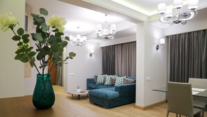 Uma sala de estar moderna com um sofá azul, uma mesa de jantar de vidro e uma mesa de centro de madeira. Há duas luminárias de cristal e um vaso com flores sobre uma mesa de madeira.
