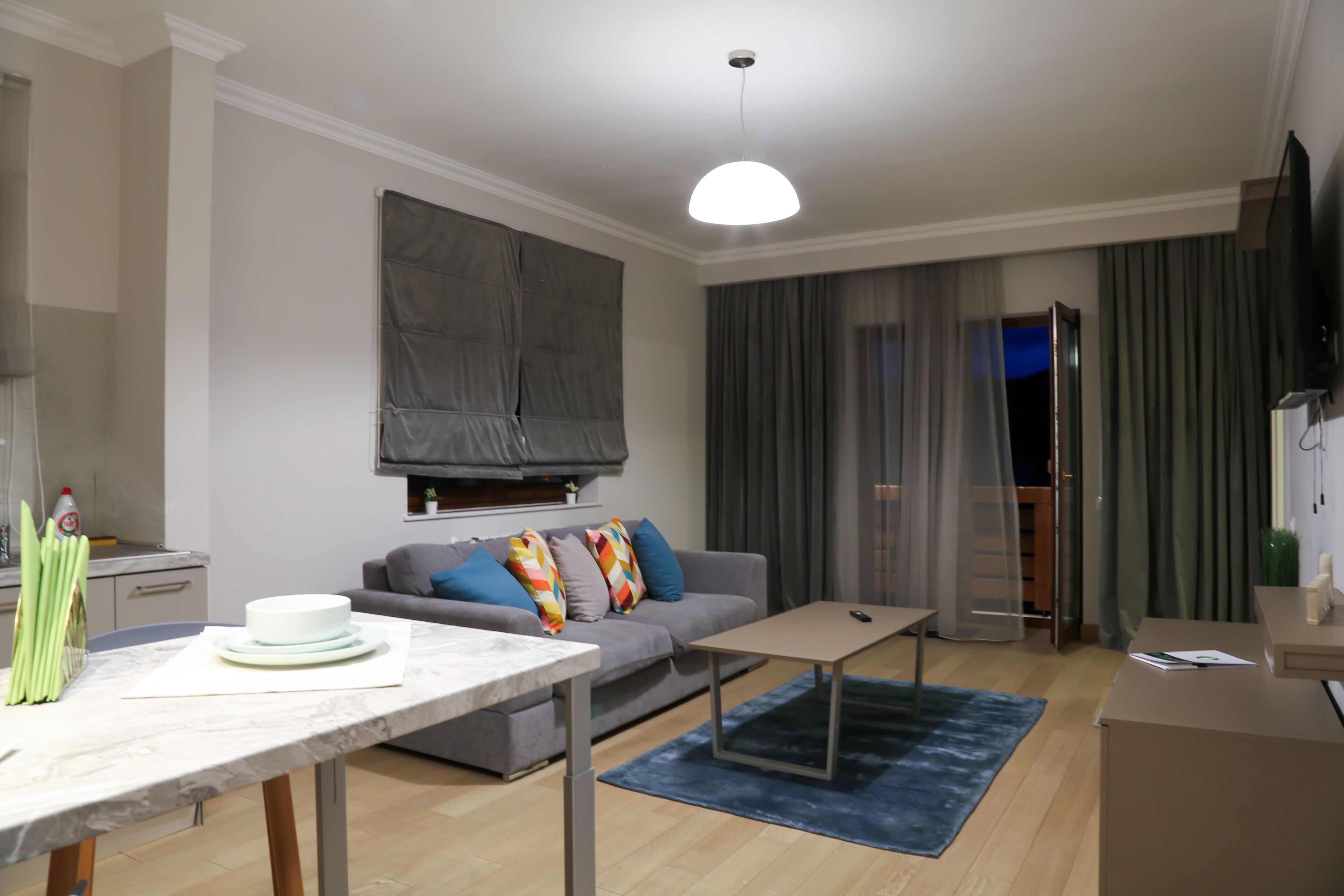 Een moderne woonkamer met een bank, een salontafel en een eettafel. Er is een televisie aan de muur bevestigd en een raam met gordijnen.