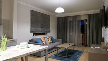 Een moderne woonkamer met een bank, een salontafel en een eettafel. Er is een televisie aan de muur bevestigd en een raam met gordijnen.