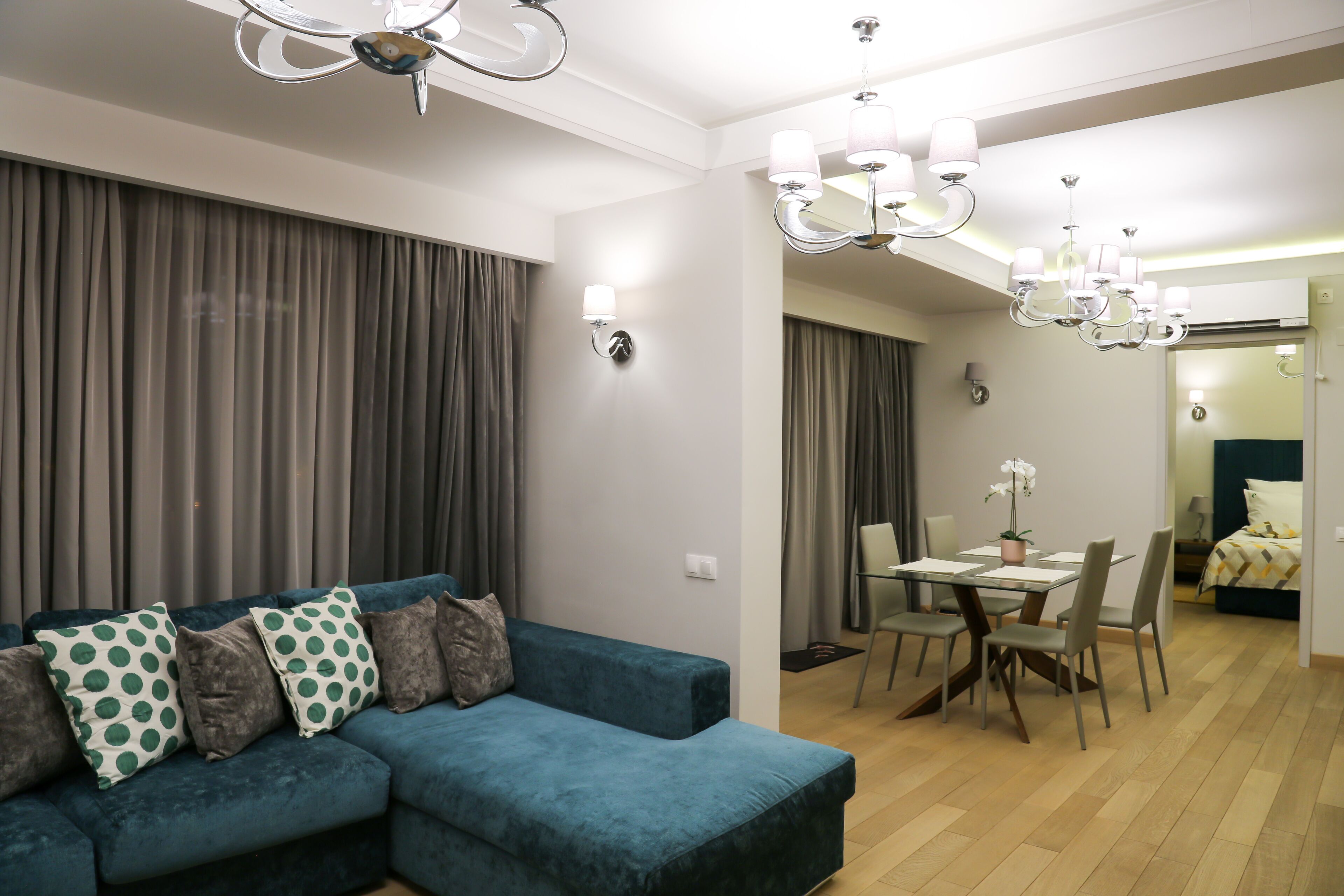 A modern living room with a teal sectional sofa, a dining area with a table and chairs, and a chandelier.