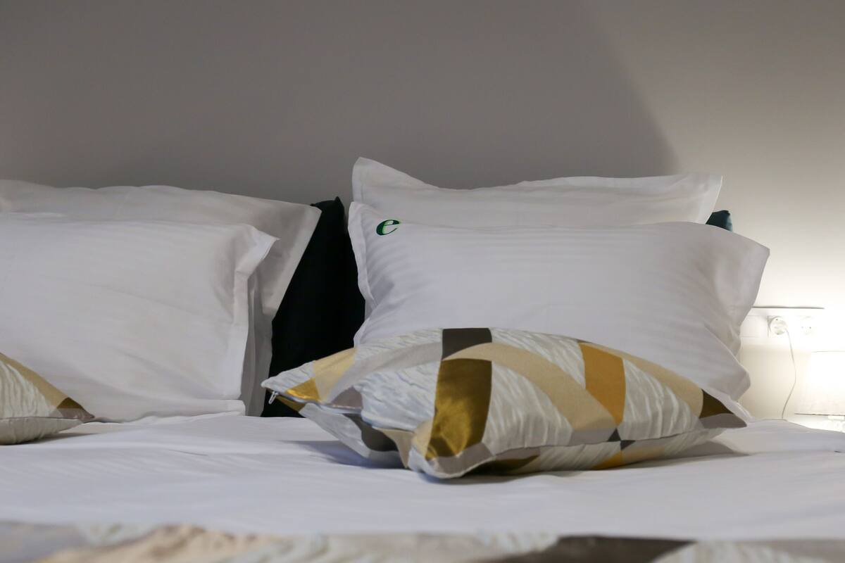 Una cama bien hecha con ropa de cama blanca y almohadas estampadas, una lámpara de noche y un reloj de pared.