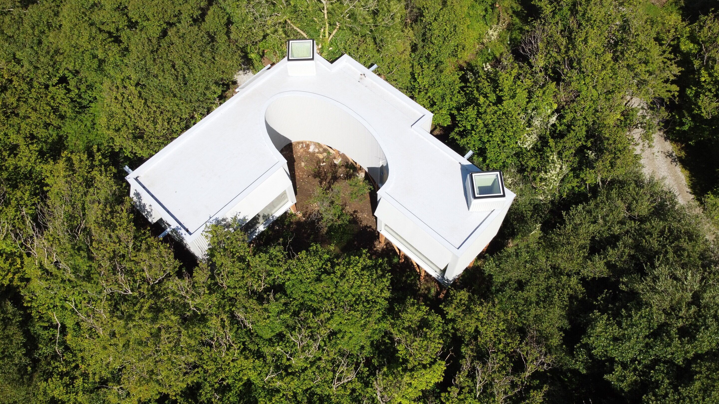 Vista aérea de una casa con una estructura de techo curva distintiva, rodeada de un bosque frondoso.
