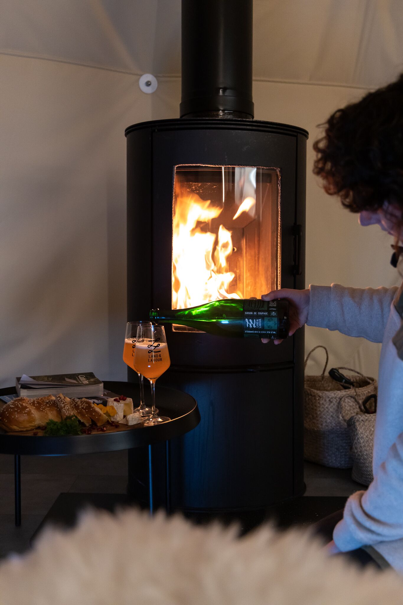 A person pouring a drink into a glass next to a wood-burning stove.