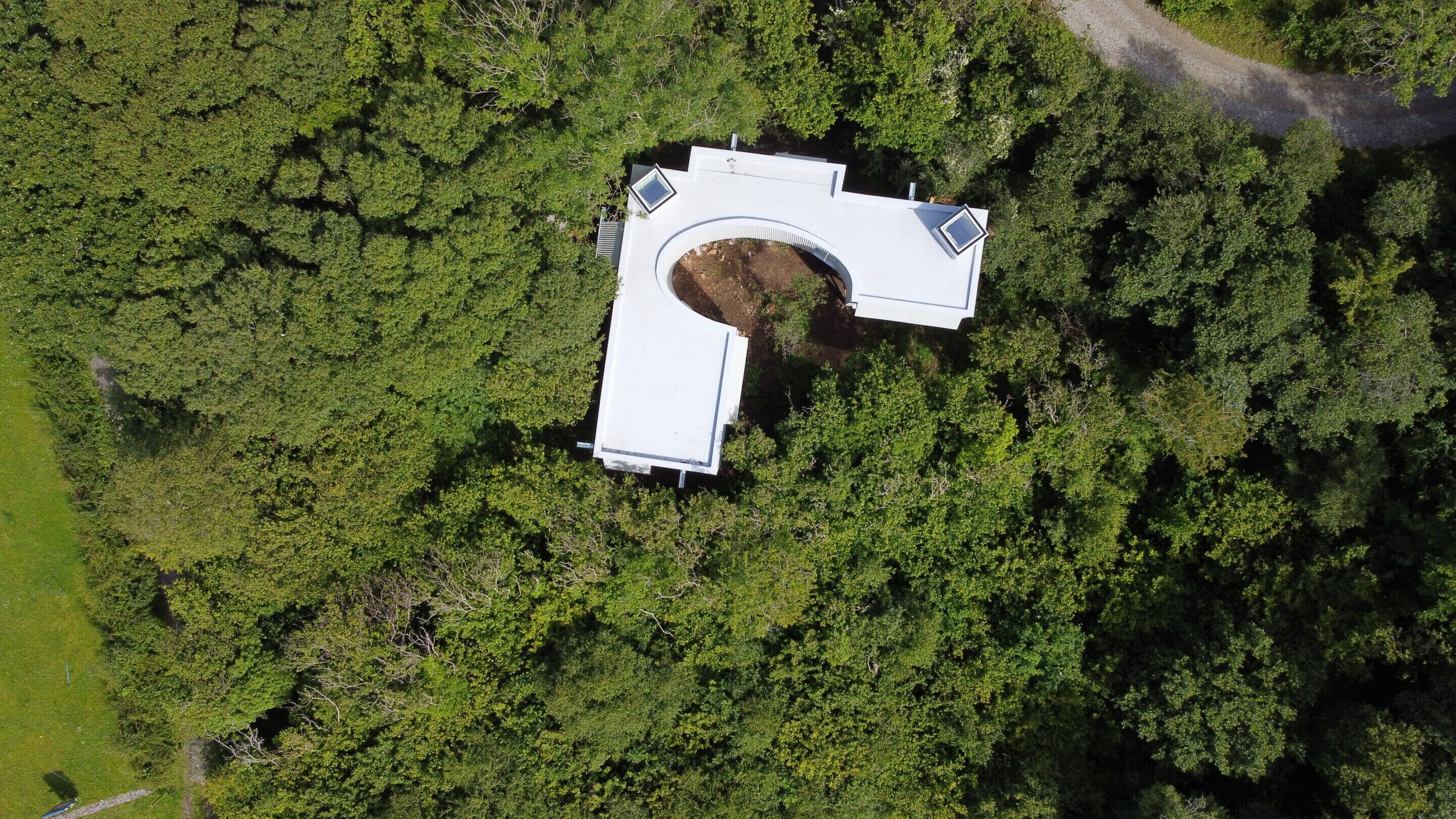 An aerial view of a building with a unique curved roof structure surrounded by dense green forest.
