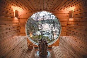 A wooden sauna with a circular window offering a view of a lake and trees. - Fernwood (Clifden)
