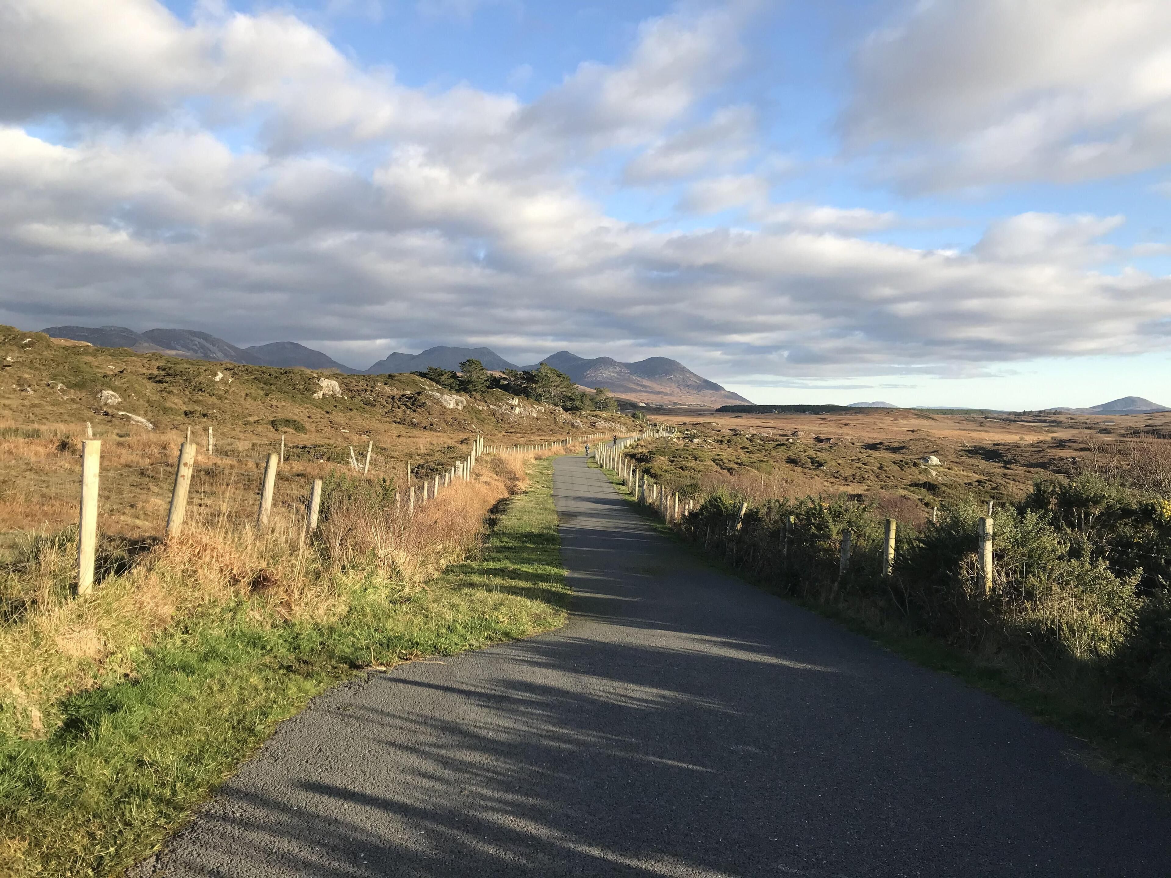 Une route de campagne bordée de clôtures en bois et d’une végétation clairsemée, qui mène vers des montagnes lointaines sous un ciel partiellement nuageux.