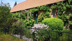 Un bâtiment en briques surmonté d’un toit en tuiles, agrémenté d’une pergola en bois et entouré d’une végétation luxuriante, notamment de plantes à fleurs.
