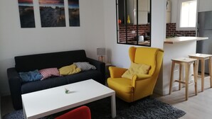 A living room with a black sofa, a yellow armchair, a white coffee table, and a red chair. There are three framed pictures on the wall, a window, and a kitchen area visible through a glass partition. - 100 m from the beach in a pretty family seaside resort (Luc-sur-Mer)