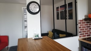 A modern living room with a wooden table, a wall clock, a sliding glass door, a red sofa, and a brick fireplace. - 100 m from the beach in a pretty family seaside resort (Luc-sur-Mer)