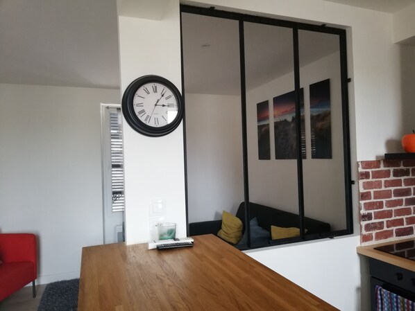 A modern living room with a wooden table, a wall clock, a sliding glass door, a red sofa, and a brick fireplace.