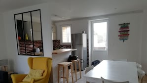 A compact living space with a yellow armchair, a white dining table, and a kitchen area with a refrigerator and a window. - 100 m from the beach in a pretty family seaside resort (Luc-sur-Mer)