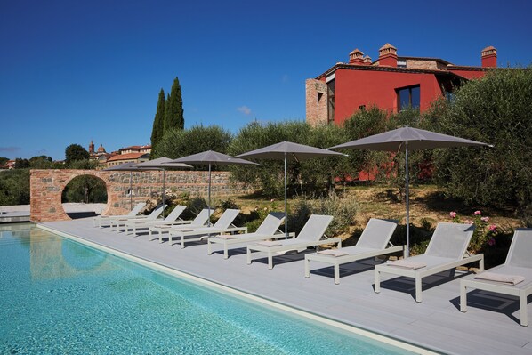 A poolside area with lounge chairs and umbrellas, a stone bridge, and a red building in the background. - Borgo 69 (Foiano della Chiana)