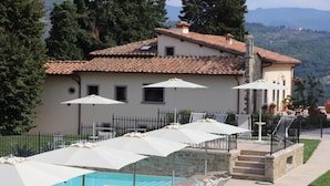 Uma área junto à piscina com espreguiçadeiras e guarda-sóis, uma escadaria de pedra e um edifício com telhado em azulejos.