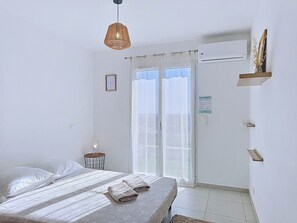 A bedroom with a bed, a hanging light, a window with curtains, a door, and a wall-mounted shelf.