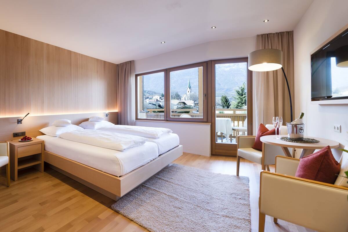 A modern hotel room with a large bed, a desk, a chair, and a view of the city through large windows.