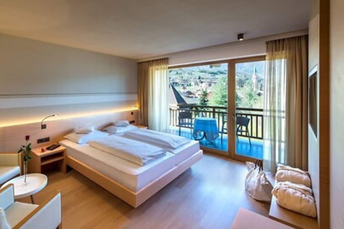 A hotel room with a large bed, a sitting area with a chair and a small table, a balcony with a view of the town.