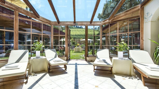 Un salón con ventanales iluminado por el sol, con varios sillones, una mesa central y vistas a un jardín exuberante y una colina.