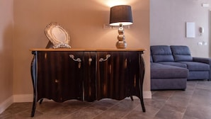 Una consola de madera oscura con un espejo y una lámpara, ubicada en una habitación con un sofá gris y piso de baldosas.