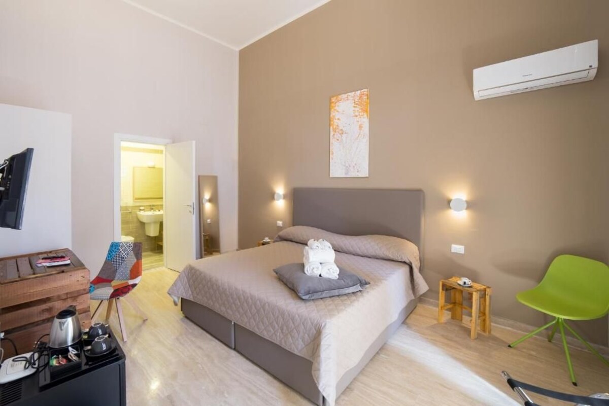 A modern hotel room with a bed, a small wooden side table, a green chair, and a kitchenette area.
