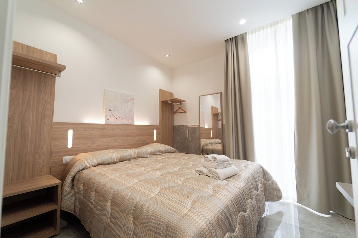 Ein Hotelzimmer mit einem großen Bett, einem Holz-Kopfteil, einem Nachttisch, einem Spiegel und einem Fenster mit Vorhängen.