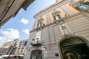Un edificio histórico con un gran arco de entrada, un balcón con plantas en macetas y un cielo azul despejado al fondo.
